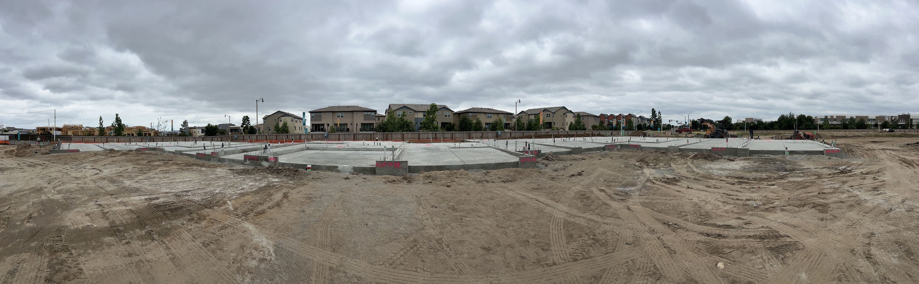 Wide panorama of poured concrete foundations across a residential lot, with finished homes in the background
