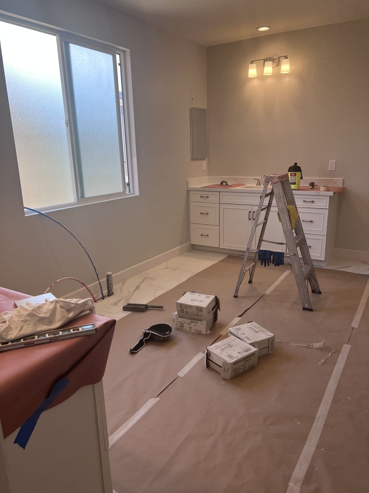 A bathroom mid-finish with a stepladder, fixture boxes, and floor protection still in place
