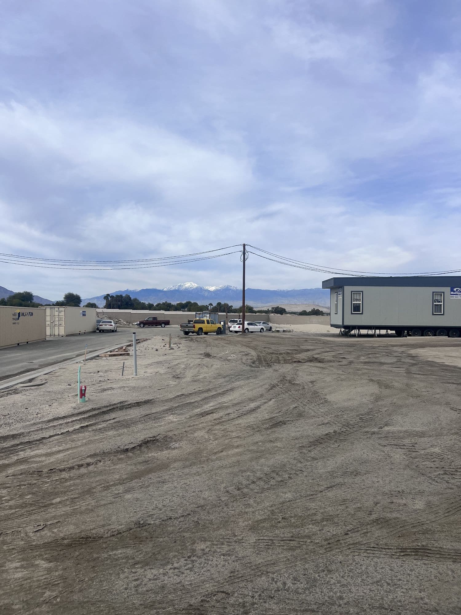 A graded dirt lot with a construction office trailer and shipping containers, mountains behind